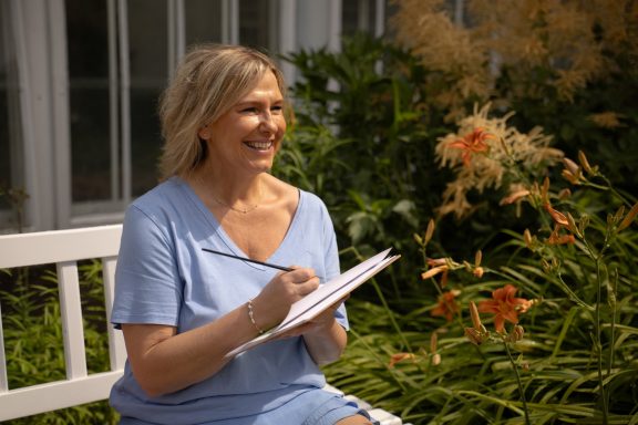 Eine lachende Frau sitzt auf einer Bank vor Blüten im Hintergrund und hält einen Skizzenblock in der Hand.