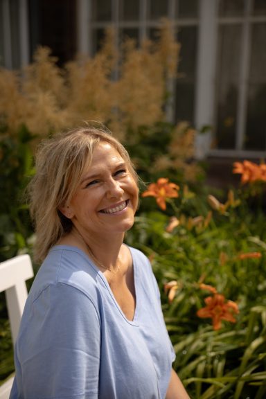 Eine Frau lacht in die Kamera sitzt vor orangenen Blüten auf einer Bank.